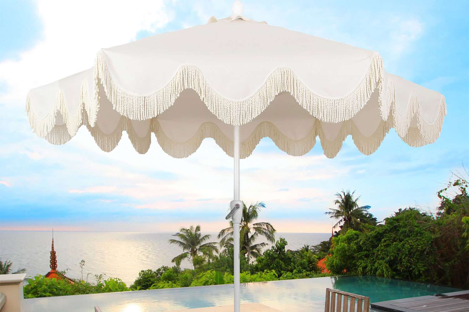 A classic cottagestyle patio umbrella with tassels set outdoors overlooking a pool tropical trees and the ocean during sunset
